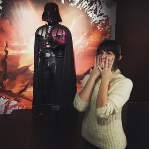 A young woman making a delighted face, standing in front of the costume of Darth Vader, from the Star Wars films.
